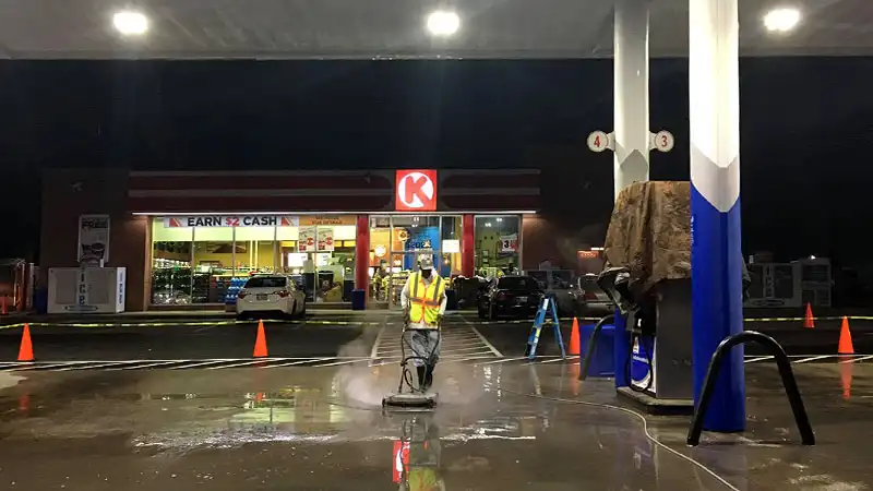 A HurriClean pressure washing technician operates a concrete surface cleaner under the brightly lit canopy of a gas station in the Louisville KY area.