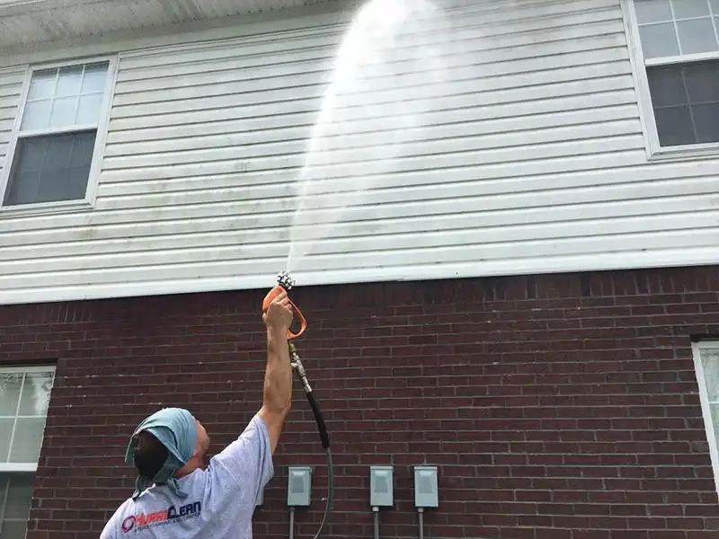 A technician wearing a HurriClean company shirt sprays Soft Washing cleaning solution on the side of a residential brick and vinyl siding home covered in algae growth in the Louisville area.
