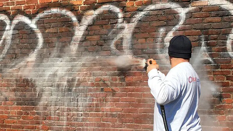 A HurriClean Graffiti Removal technician wearing a grey company shirt and black skull cap power washing a building's brick wall in the Crestwood, KY area.