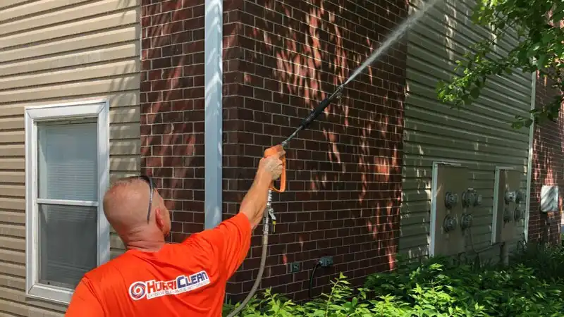 A HurriClean wash tech in a bright orange uniform shirt using a Soft Wash spray wand to clean a vinyl siding and brick exterior property exterior in the Crestwood, KY area.