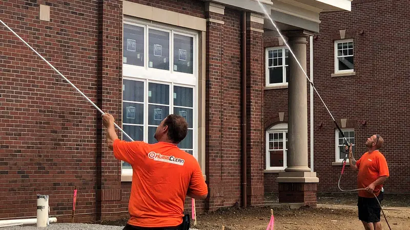 A pair of HurriClean workers wearing orange company uniforms using long Soft Washing wands to clean a commerical building's brick facade in the Goshen, KY area.
