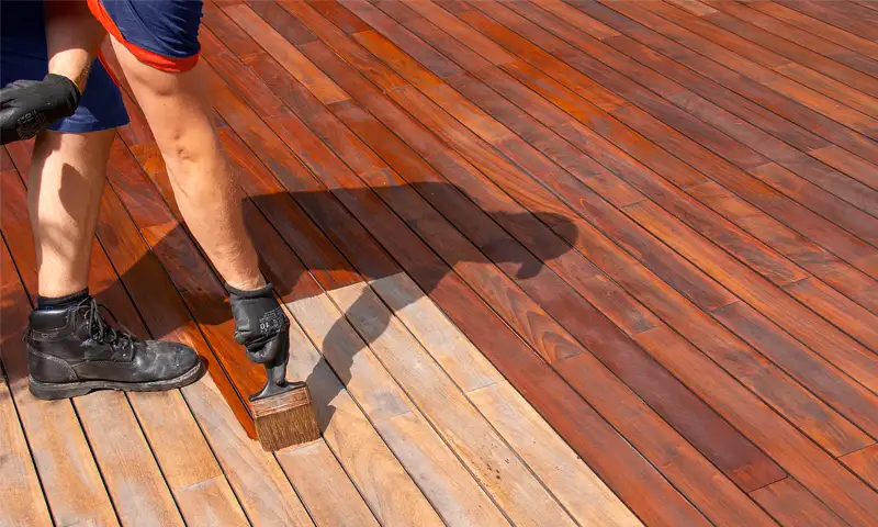 A HurriClean Deck Restoration technician is partially visible applying redwood colored stain to a wooden deck at a job site in the Louisville KY area.